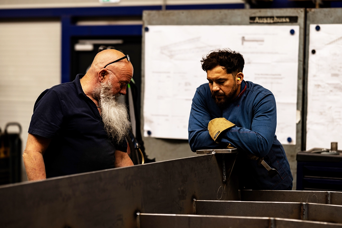Zwei Mitarbeiter besprechen sich in der Halle über einem Stahlbauteil, im Hintergrund hängen Zeichnungen